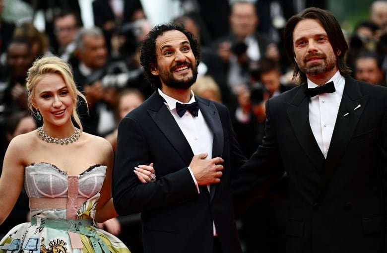 Three people in formal outfits pose next to one another. Behind them is a crowd of photographers.