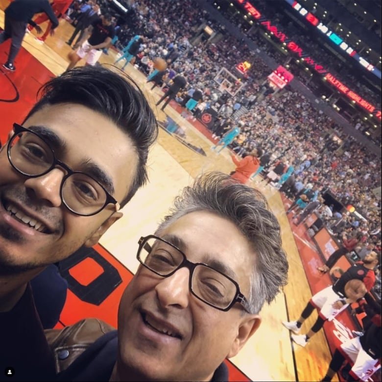 Two men, both wearing glasses, smile at the camera with a basketball court, a crowd, and two players in the background.