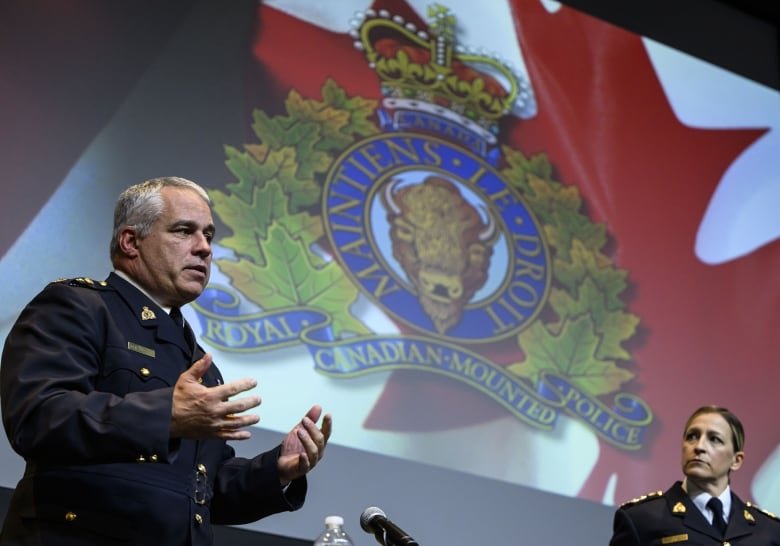 A police officer is pictured with the RCMP logo in the background on a screen as another police officer looks on.