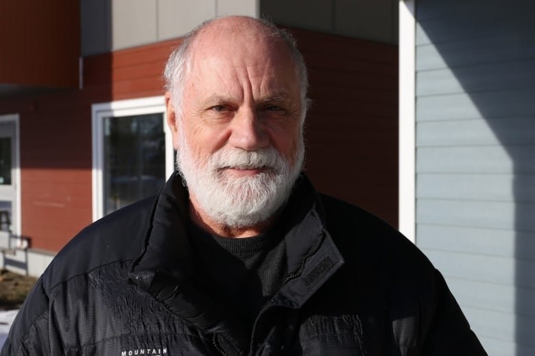 A smiling, mostly bald man with a white beard smiles at the camera. He is wearing a black ski jacket and stands in front of a grey and rust-coloured building.