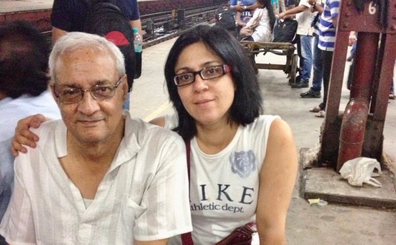 An aged south Asian man and a south Asian woman sit on a bench on a busy train platform. The woman has her right arm around the man.