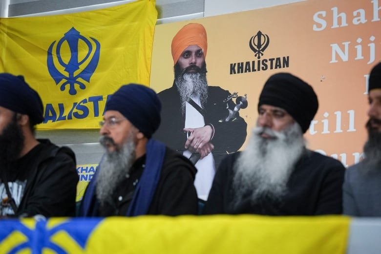 A poster of a Sikh man holding a ceremonial sword is pictured behind a row of Sikh men sitting at a table.