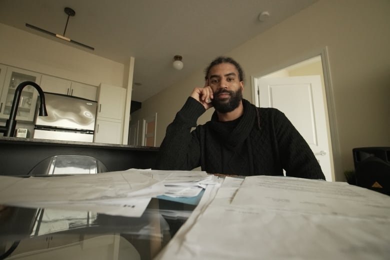 A man wearing a dark sweater sits at a table with a large pile of papers in front of him. Behind him is the kitchen. He is not smiling.