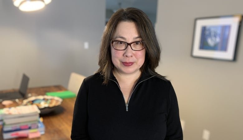 A woman with black hair and a black zip sweater sits on the end of a wooden table. A stack of books and a laptop are behind her.