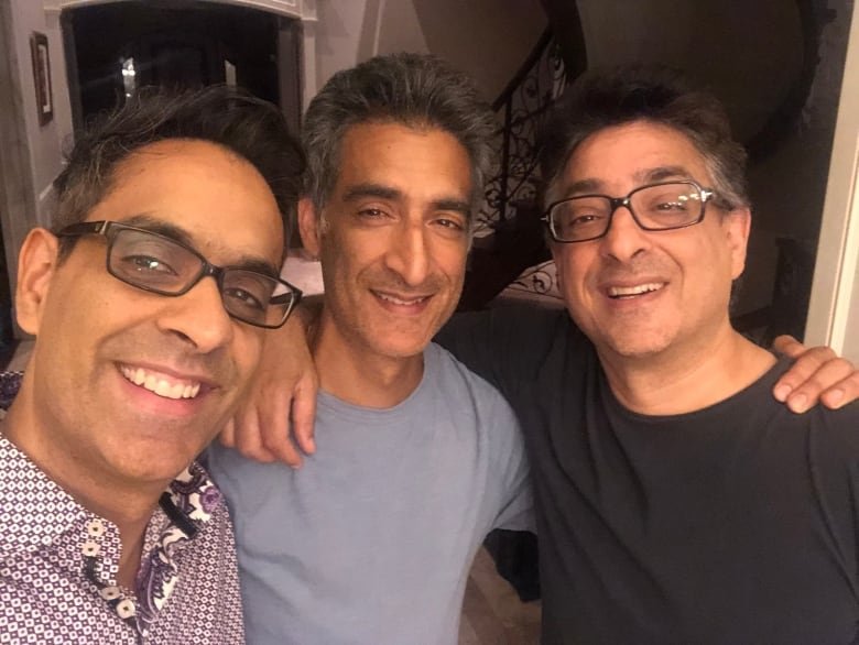 Three men smile widely at the camera. The ones on the left and right are wearing glasses while the one in the middle is not. The man on the left wears a patterned button-up shirt, the middle man wears a blue-grey t-shirt, and the man on the right wears a dark grey t-shirt.