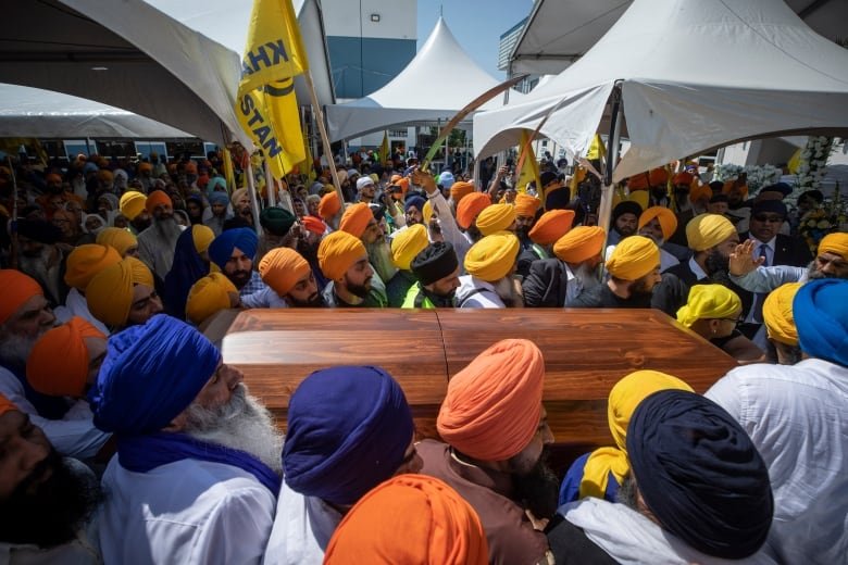 A brown wooden casket is carried through a dense crowd at a funeral. A yellow flag is seen in the background.