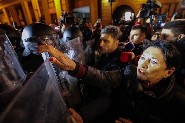 An older woman with dark hair reaches with her hand toward a police officer in riot gear and shield in a nighttime photo. In the background other protesters are face-to-face with officers.