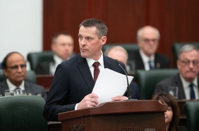 A man wearing a suit stands at a podium. He holds a piece of paper. He looks off.