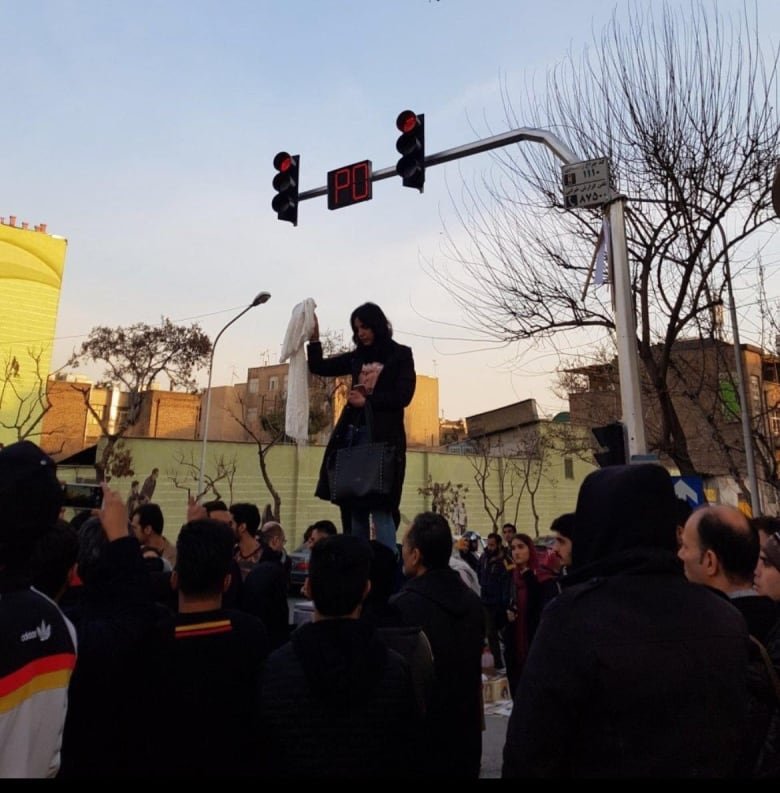 A woman in a black jacket stands on an electrical box holding her white hijab in her hand, above her head. A crowd of people stands around her.