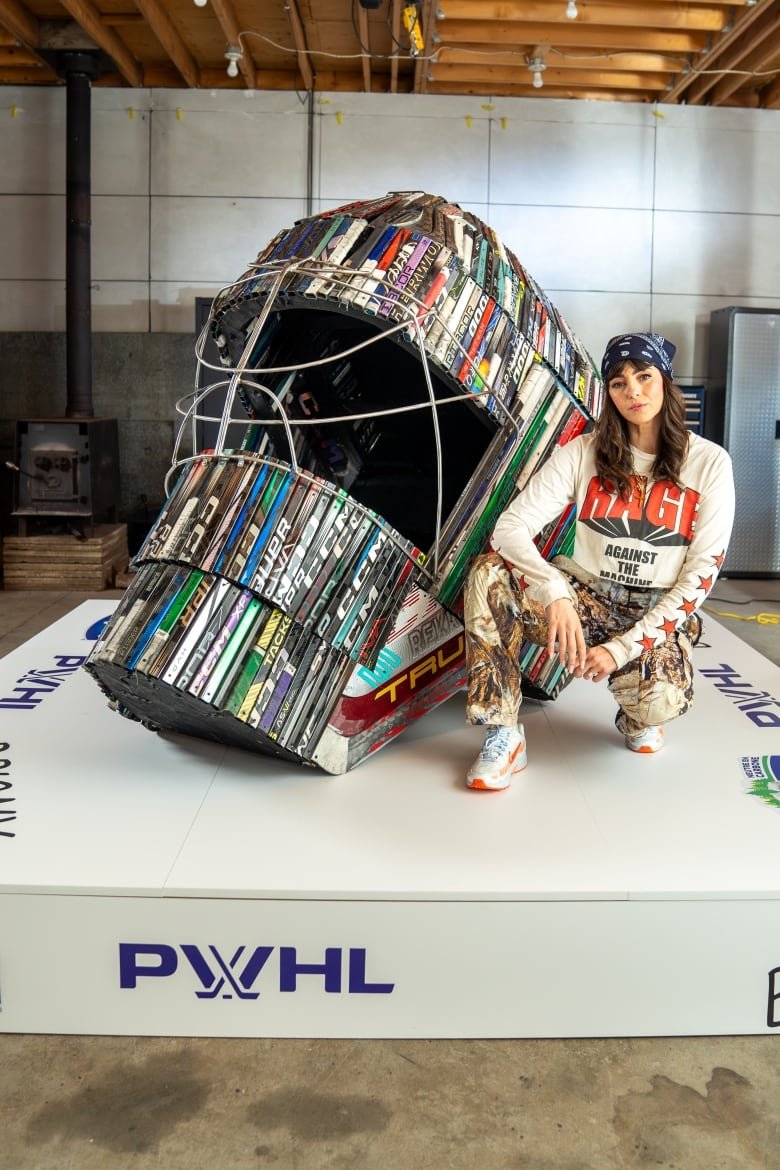 Briony Douglas (30s, dark hair, white Rage Against the Machine T-shirt, blue bandana) squats down next to an enormous goalie mask.