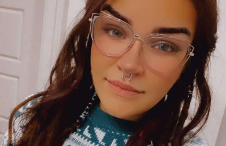 Young woman with long dark hair in dreadlocks, wearing glasses, a nose ring and a green and white sweater.