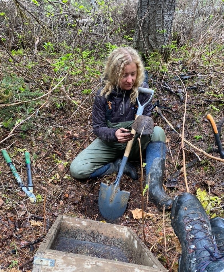 A woman looks at something small in her hands while cradling a blue shovel.