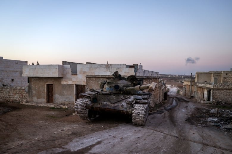 A tank is shown in front of some low-rise concrete buildings.