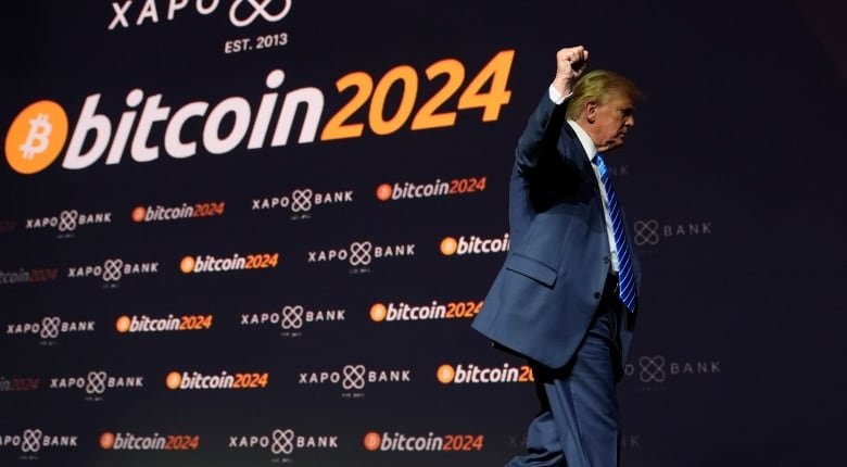 A man raises his fist as he walks off stage. The photo is taken from slightly below, and the "bitcoin 2024" lettering on the black backdrop behind the man is slanted.