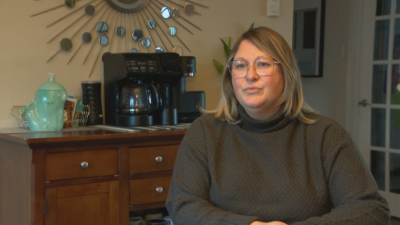 A woman wearing glasses sit inside her home with furniture and a coffee machine in the background.