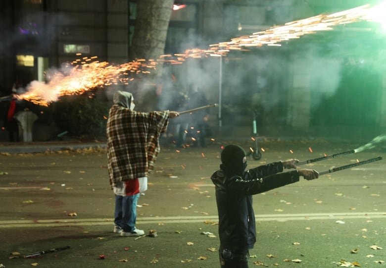 Two people, one hooded and the other wearing a dark ski mask, hold flares and shoot them on a city street in a nighttime scene.