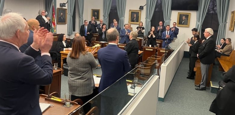 About two dozen people in business attire stand and applaud an older gentleman at the right of the photo.