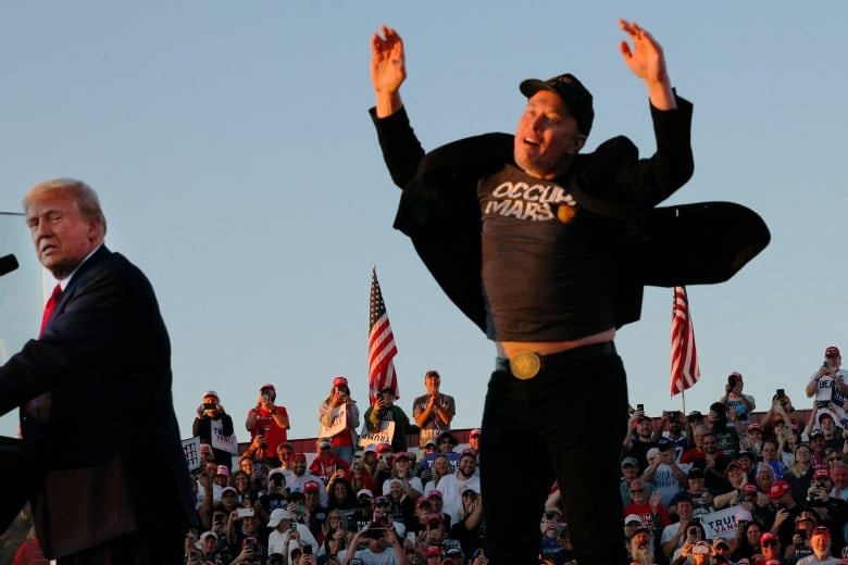 A man in a black ball cap and dark clothing jumps into the air so his t-shirt rides up and shows his stomach as an older blond man to his left stands at a microphone and looks over his shoulder at the scene.