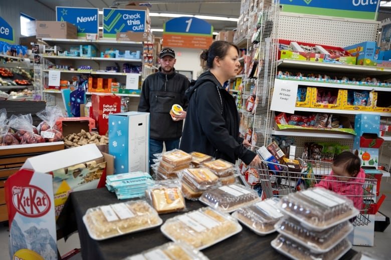 An inside view of a grocery store with shelves lined with food. A mother is pushing a cart with a baby inside and a man is holding a tu.