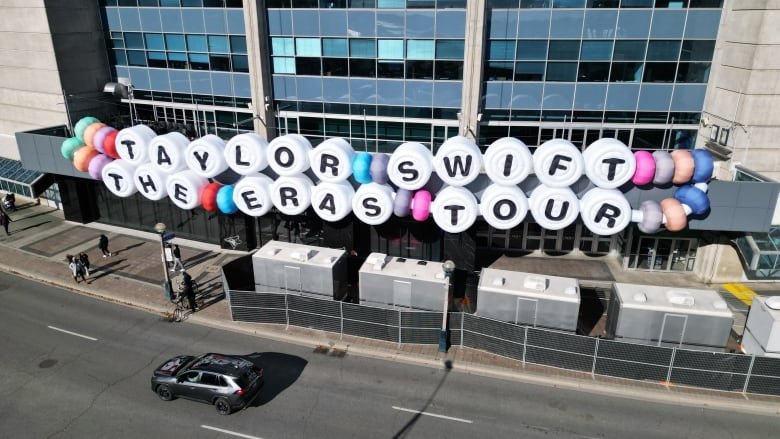 A giant white friendship bracelet spells out 'Taylor Swift The Eras Tour' one letter at a time across the front of a stadium.