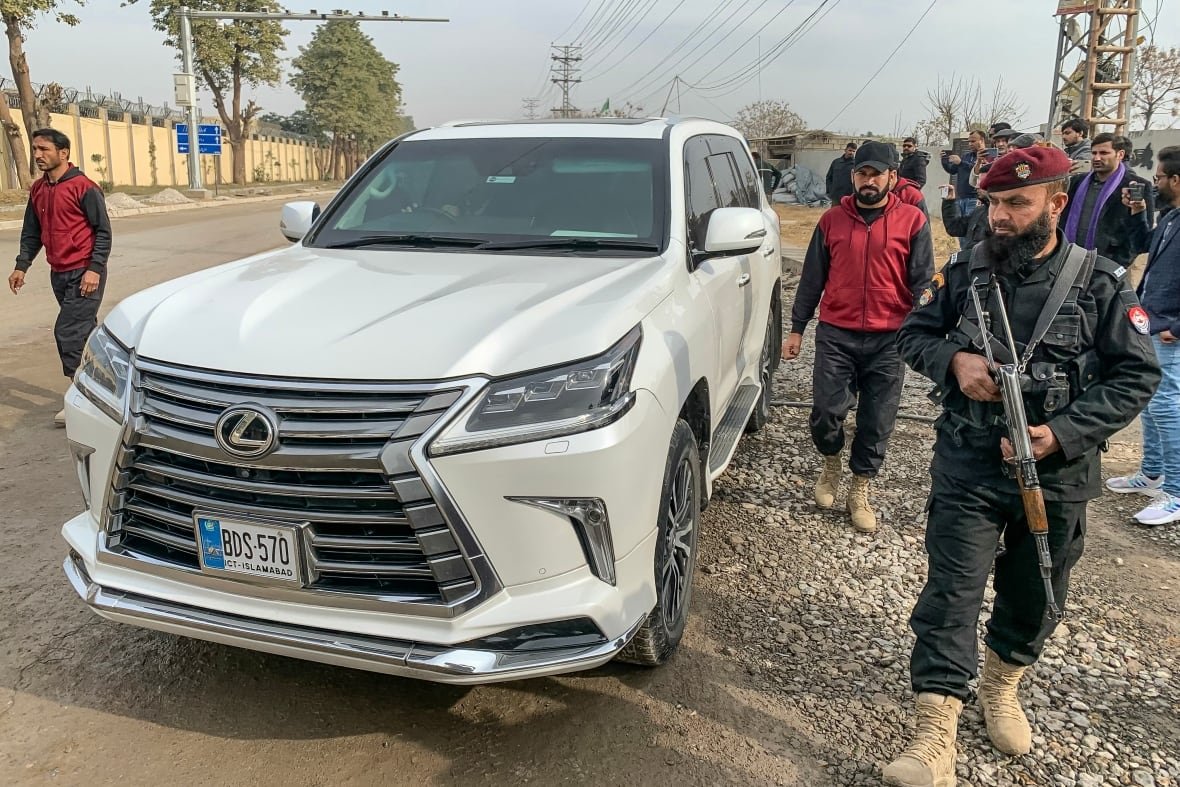 Several men, one with a long gun, are shown standing around a white vehicle.