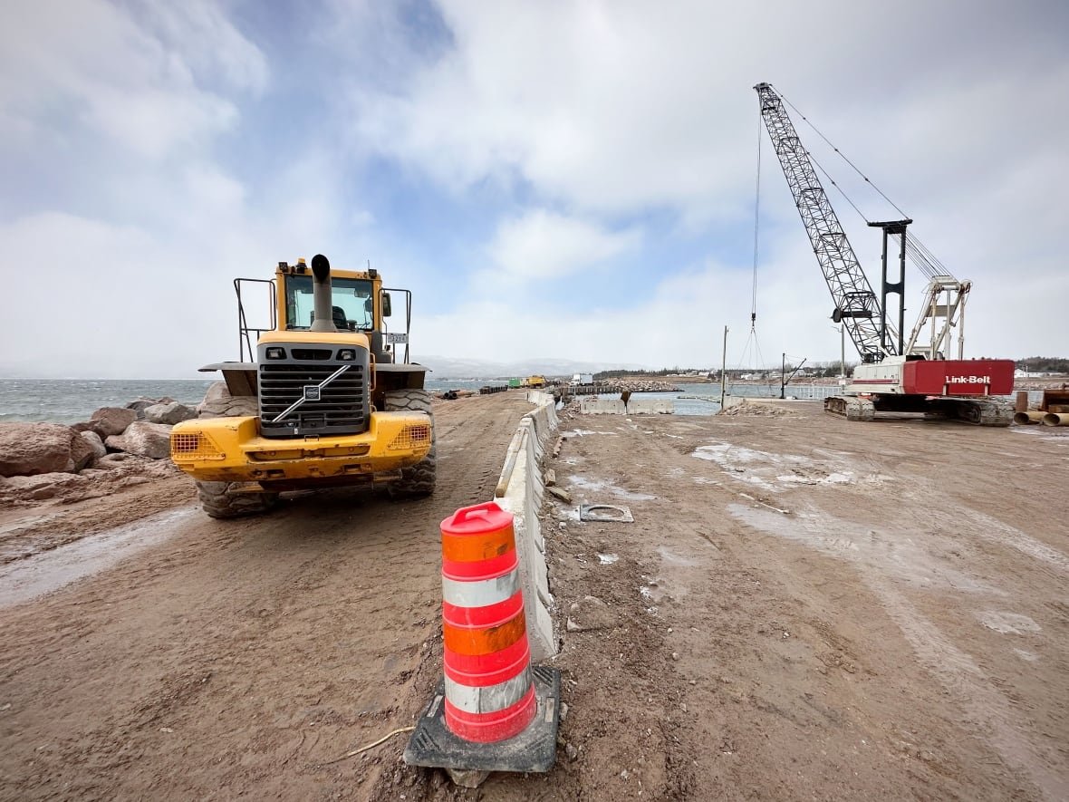 Wharf under construction with a large yellow tractor on the left and a large red and white crane on the right with a pink fluorescent traffic cone in the middle.