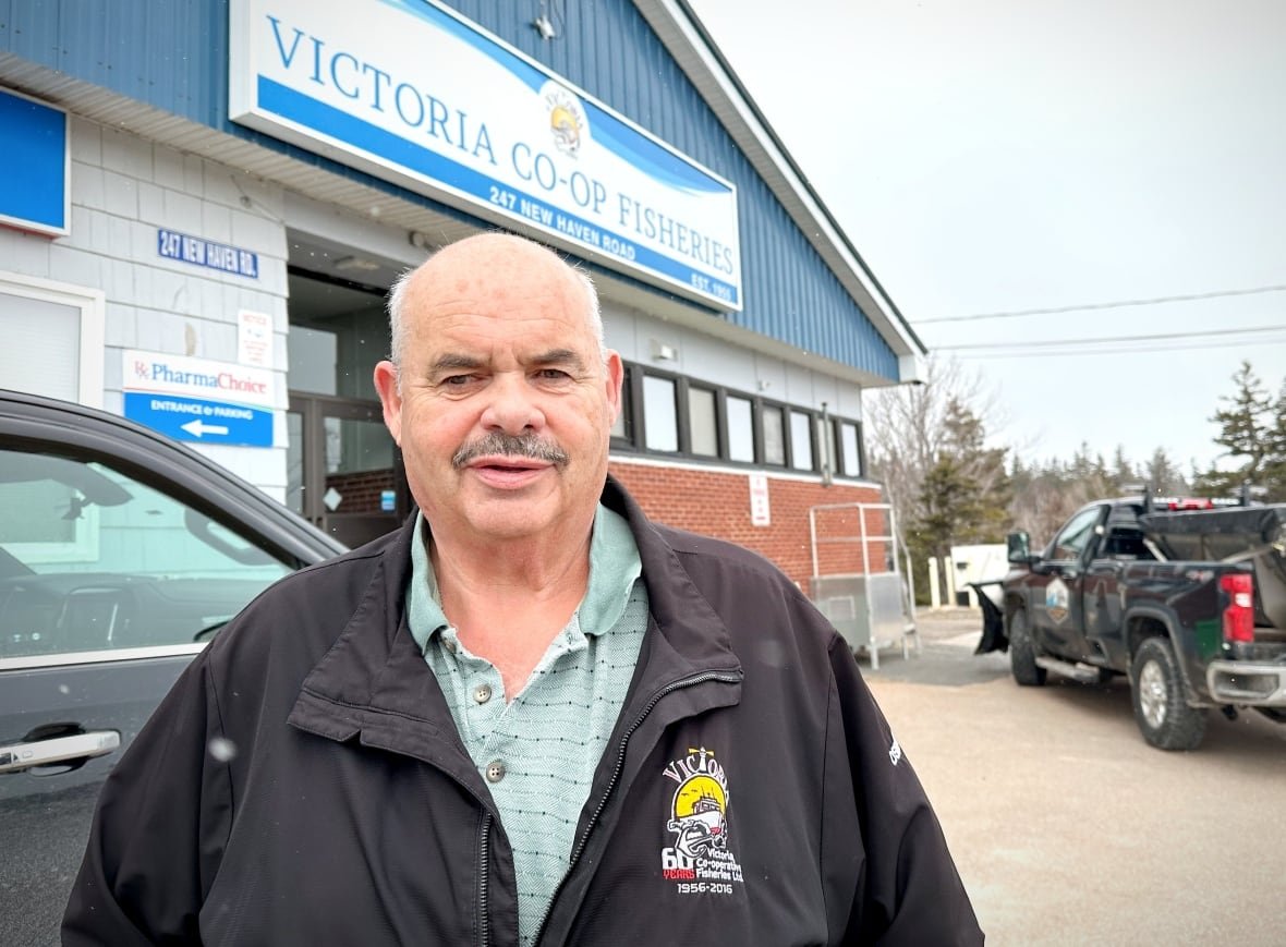 A balding man with white hair, dark grey moustache wearing a green shirt and a black jacket stands in front of a fisheries building.