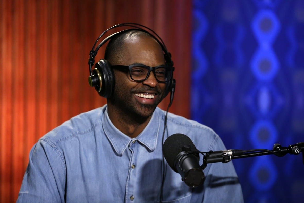 A smiling man wearing thick-rimmed glasses and over-ear headphones sits in front of a studio microphone.