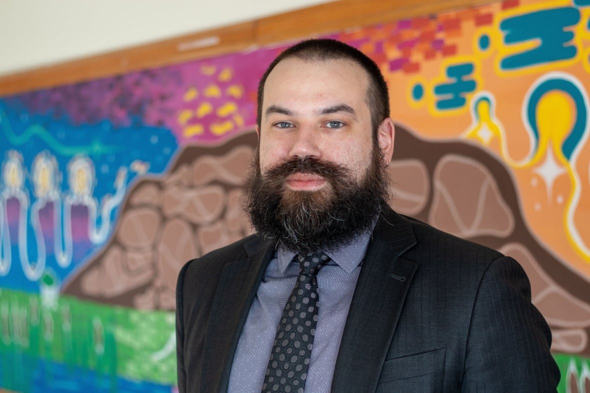 daniel-cox A person wearing a suit and grey shirt stands in front of a multi-colour mural.
