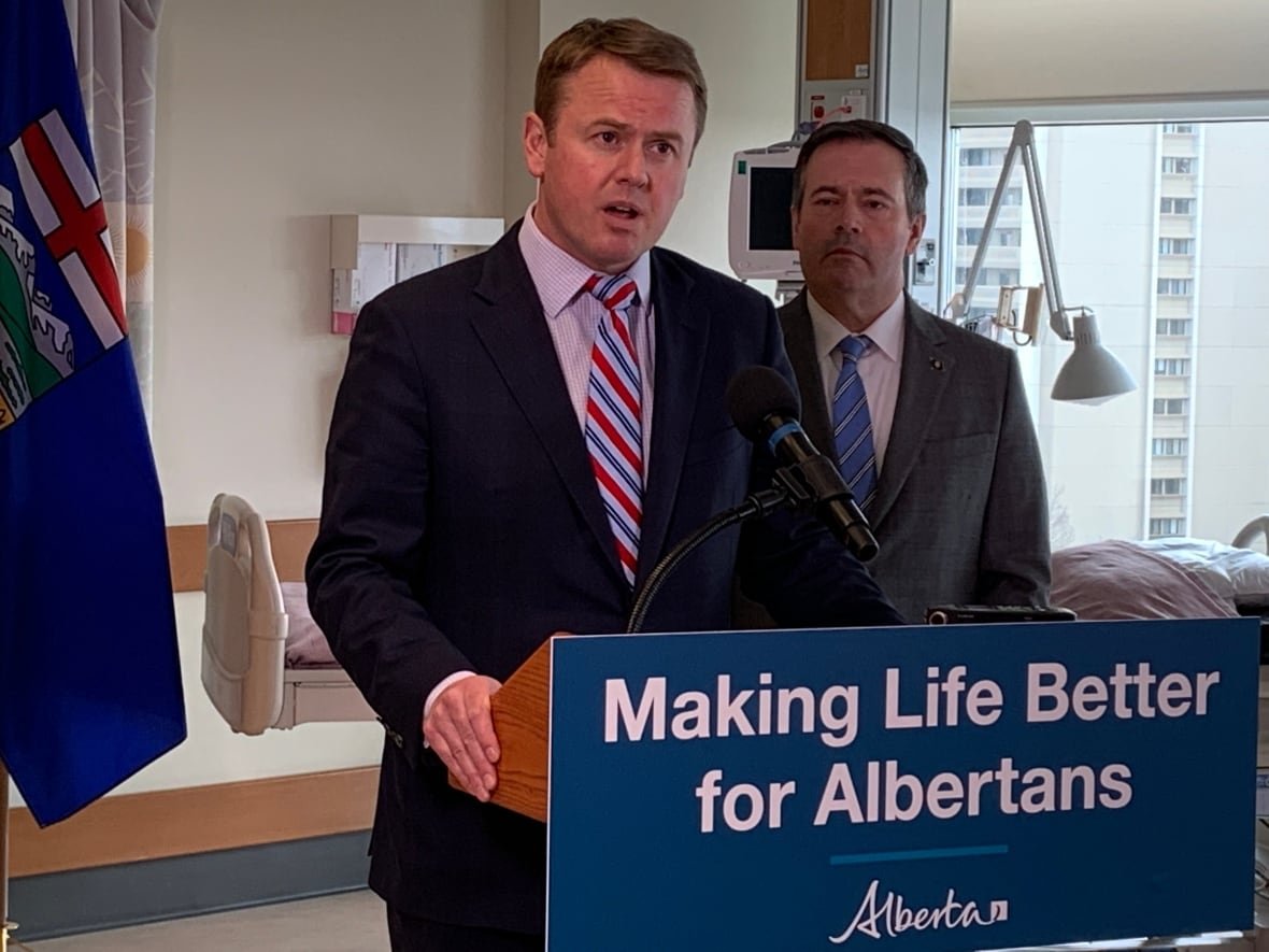 surgical-wait-times-presser-edmonton A man talks at a lectern in a hospital setting. another man watches.