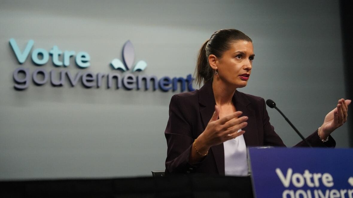 genevieve-guilbault-1 A woman with a slicked back pony tail and a burgundy blazer speaking to the media. She sits at a desk. A sign saying "Votre gouvernment" is behind her.
