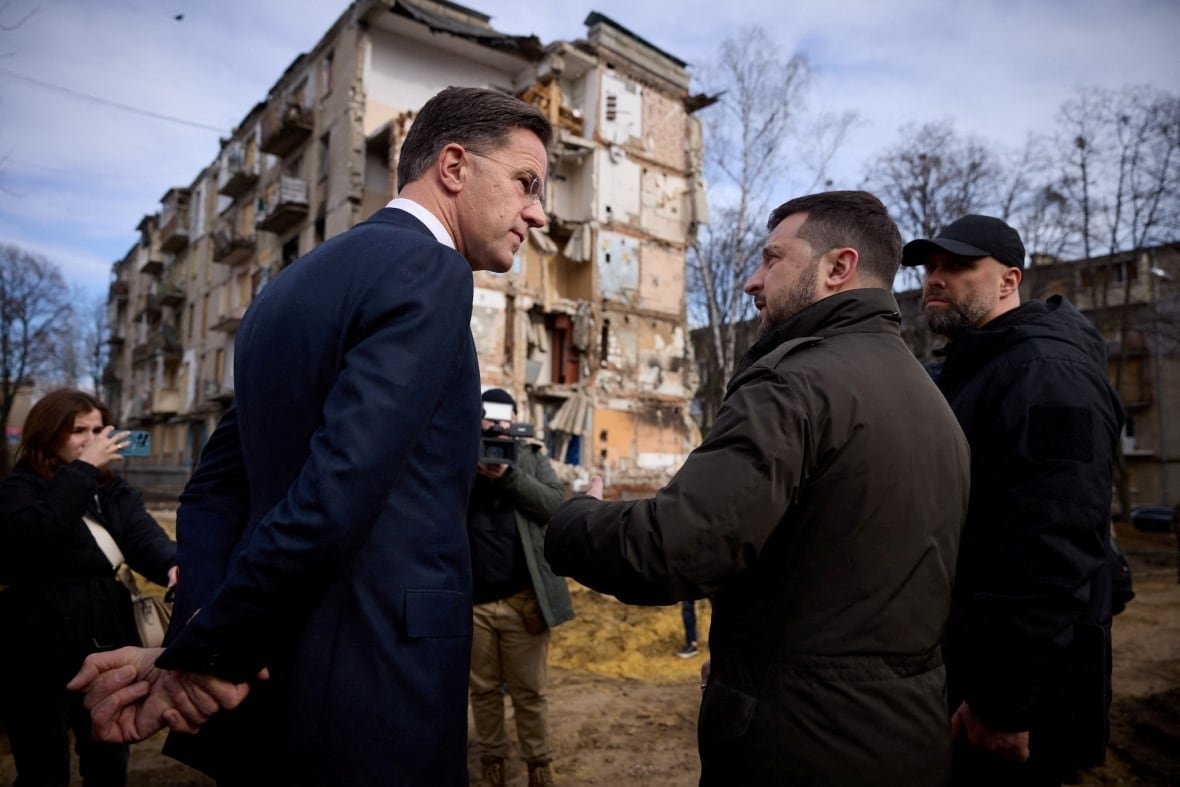 A man in a suit wearing glasses listens as a bearded man in a jacket speaks. They are shown outdoors, with a heavily damaged low-rise residential building shown in the background.