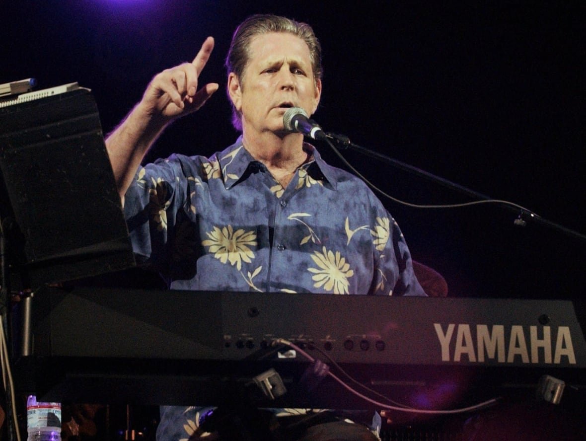 A cleanshaven man is shown onstage pointing while behind a keyboard.