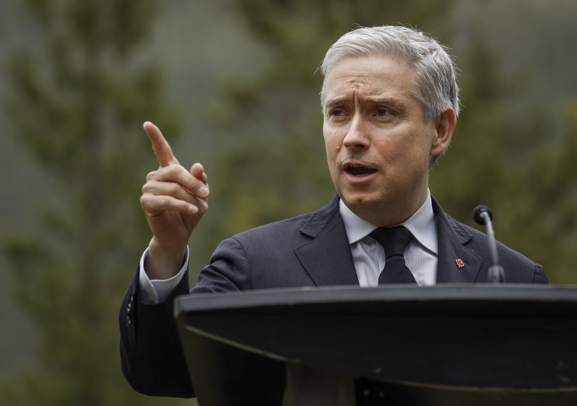 a man with grey hair and a blue suit talks at a podium while pointing with his right index finger