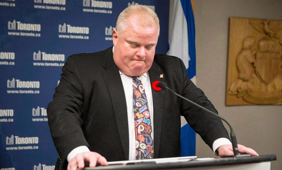 ford-crack-tie-20180705 A man in a suit braces his hands on a podium in front of a backdrop covered with the city of Toronto's logo. He has his head dipped and his mouth screwed up. He is wearing a garish tie covered in sports logos.