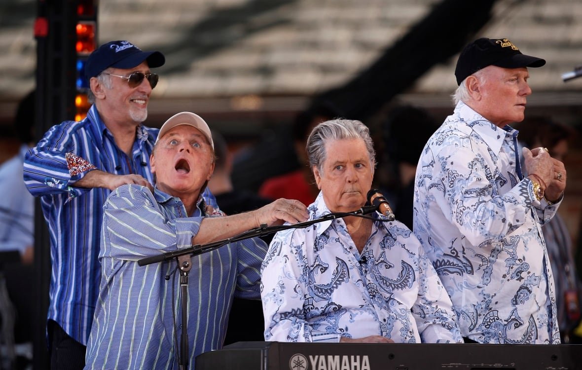 Four male musicians including three in baseball caps giving each other shoulder rubs on stage.