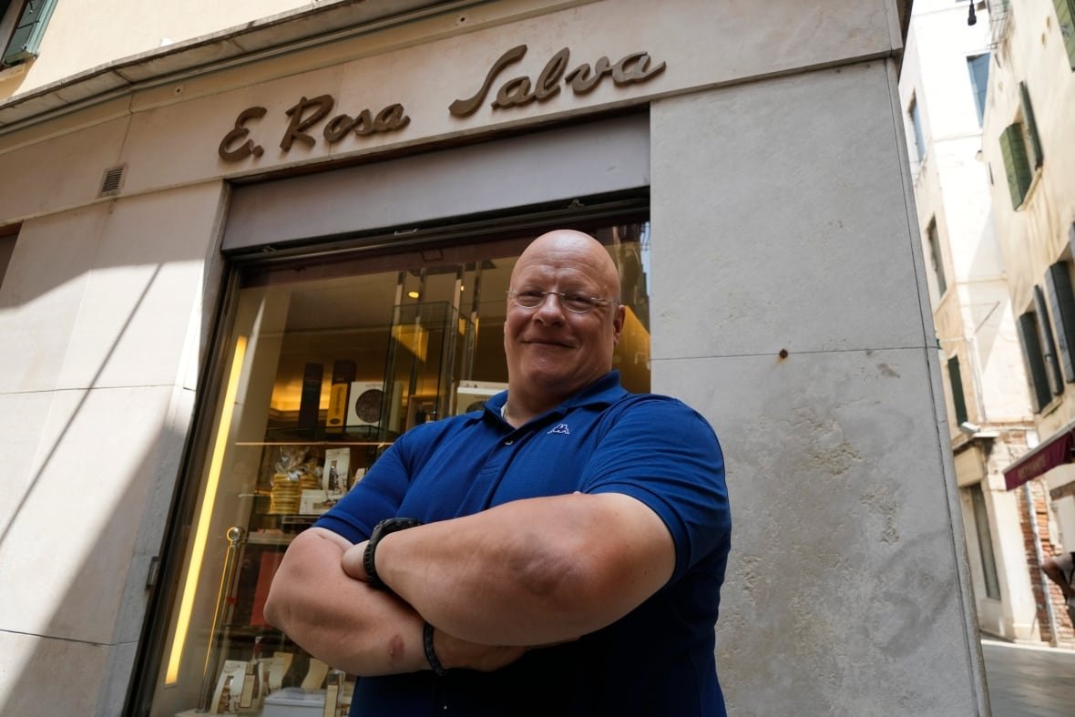 correction-italy-bezos-wedding-vendors A bald man in glasses stands with his arms folded in front of a store building with the words "E. Rosa Salva" above the door in a cursive font.