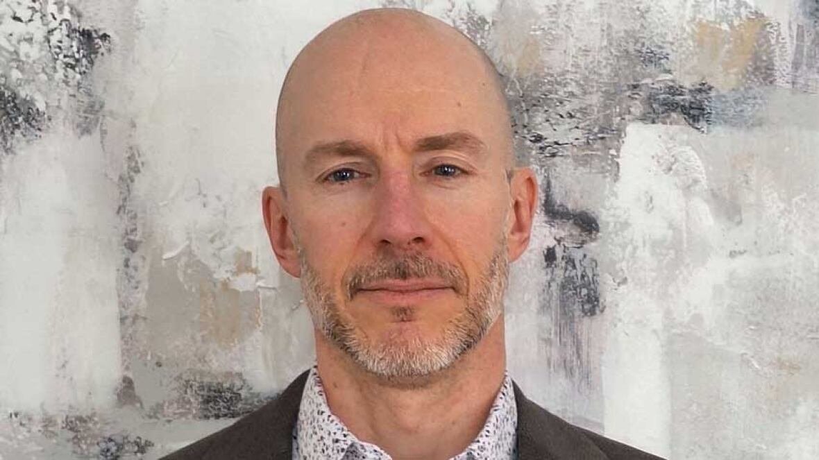 dr-yves-leger-chief-medical-officer-of-health-new-brunswick A portrait of a bald man with a salt and pepper goatee, standing in front of a wall, wearing a dark blazer and a white dress shirt with neutral-tone small flowers on it.