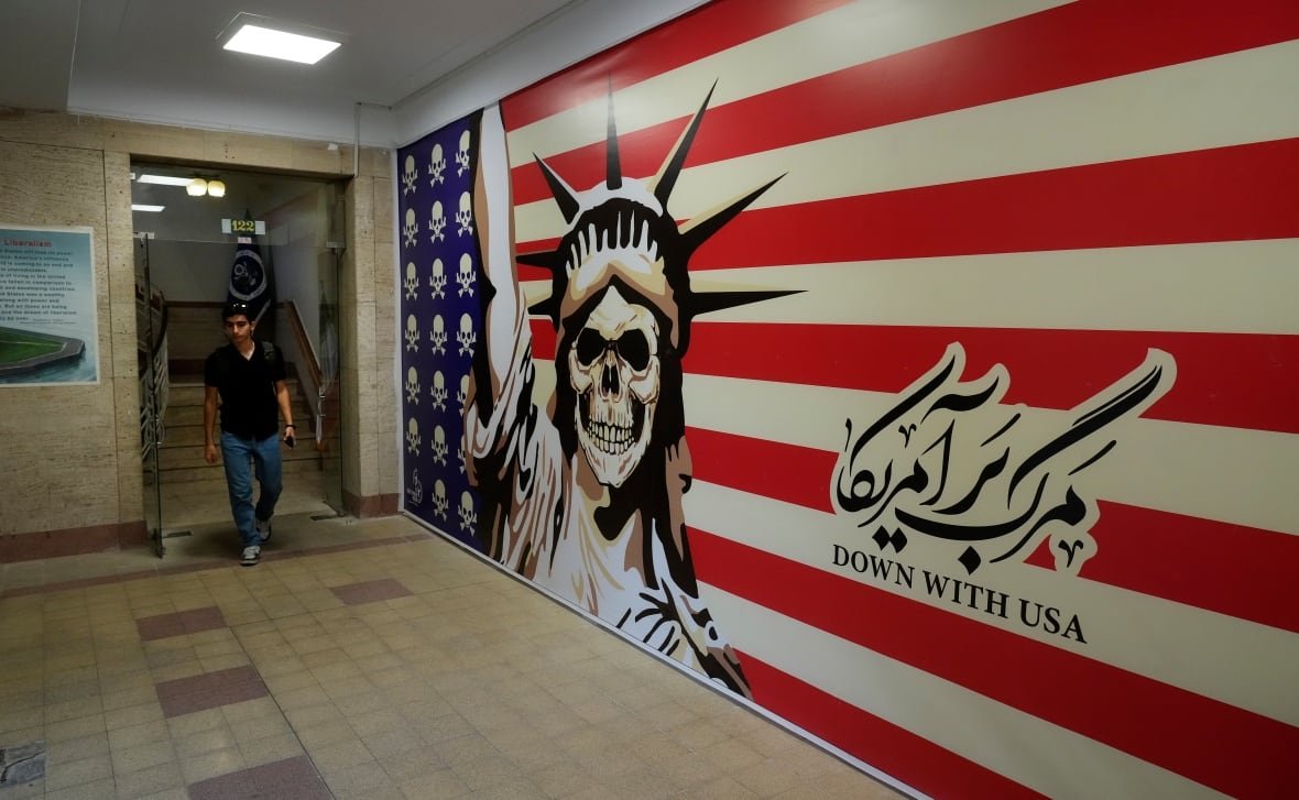 iran-1953-coup A man walks through a doorway next to a mural of a US flag with a skeleton that says 'Down with USA'