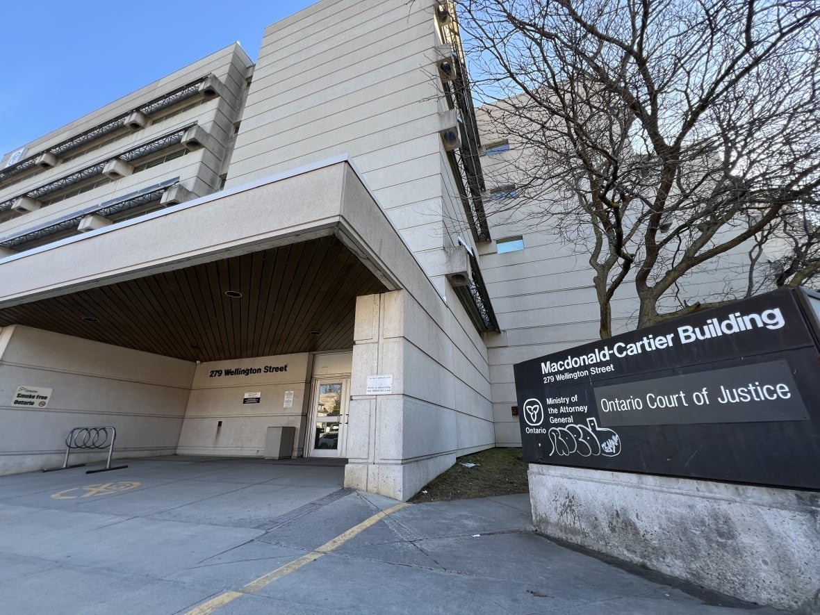 kingston-ontario-court-of-justice A sign reading "Ontario Court of Justice" is shown outside a tall concrete building. It's a clear day with a blue sky.