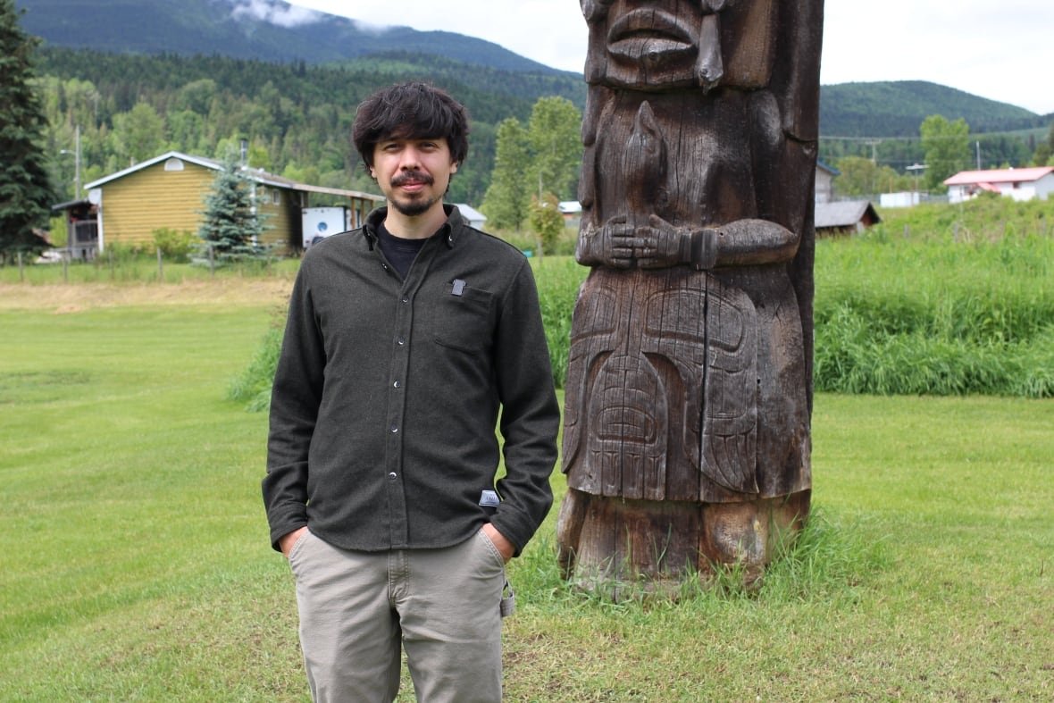 kolin-sutherland-wilson A man wearing a dark green button-down shirt and light green pants poses for a photo in front of a totem pole.