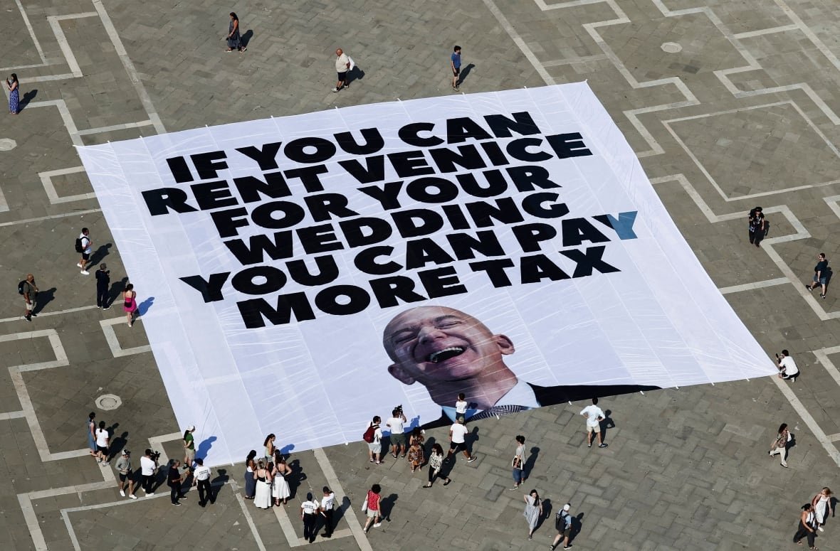 people-bezos-wedding-protest An aerial shot shows a few dozen people standing around a massive banner, unfurled and laid out on the ground. It's white with a picture of a bald man laughing, and the all-caps words: IF YOU CAN RENT VENICE FOR YOUR WEDDING YOU CAN PAY MORE TAX