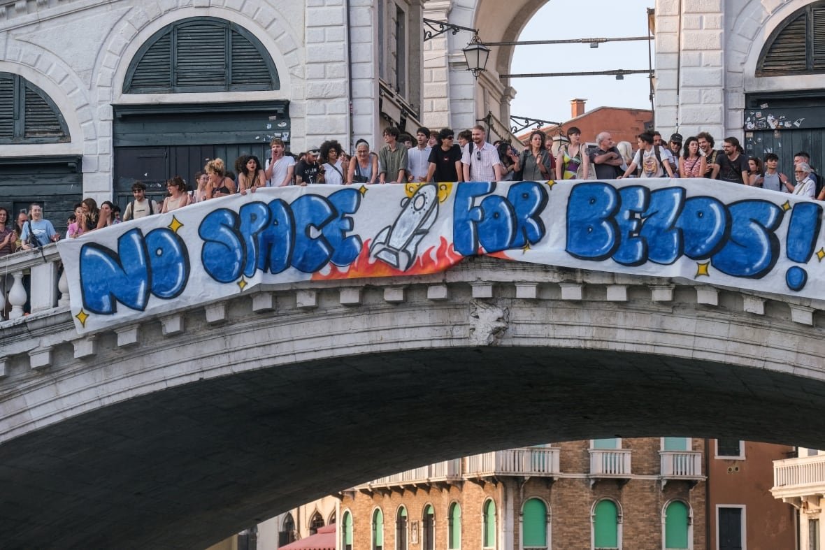 people-bezos-wedding-venice People stand on an arched stone bridge unfurling a hand-painted banner with a rocket ship and big blue bubble letters that read: "No Space for Bezos!"