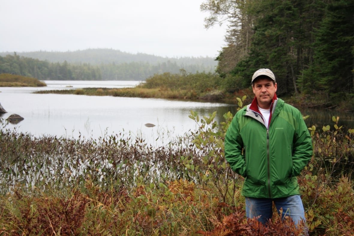 ray-plourde A white man stands in a marshy area in front of a lake wearing a cap, jeans and a green windbreaker