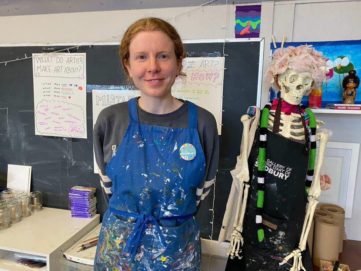 sarah-blondin A woman in a blue artist apron splattered with paint stands next to a chalk board and a skeleton in a classroom.