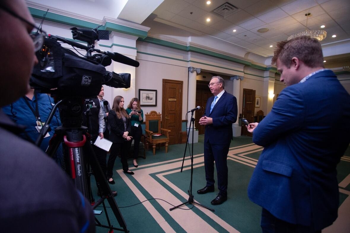 tony-wakeham-scrum A man in a blue suit speaks into a floor-based microphone, surrounded by video cameras and journalists