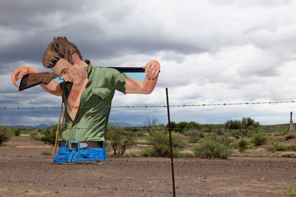 usa-dailylife A massive cutout of an illustration of James Dean next to a barbed wire fence in the desert. In the illustration, Jean bows his head, his green shirt unbuttoned halfway exposing his chest, holding a shotgun over both shoulders.