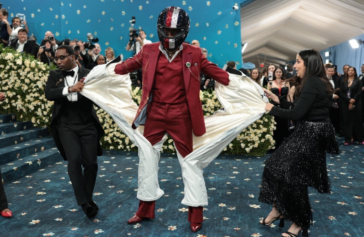 usa-met-ball-5 A person stands while two other people tear a white outfit away from him, revealing a red suit. He is also wearing a bedazzled motorcycle helmet.