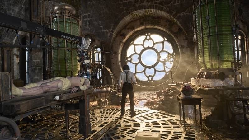 Interior shot of a fantastical laboratory in a steampunk style. A man stands in front of an enormous circular window, his back to the viewer. Behind him is all manner of old-fashioned lab equipment, very ornate in its making. Cadavers and human body parts lie on various tables and slabs.