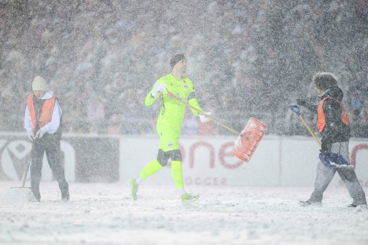 A soccer player dressed in neon yellow carries a shovel on a soccer pitch during a snowstorm.